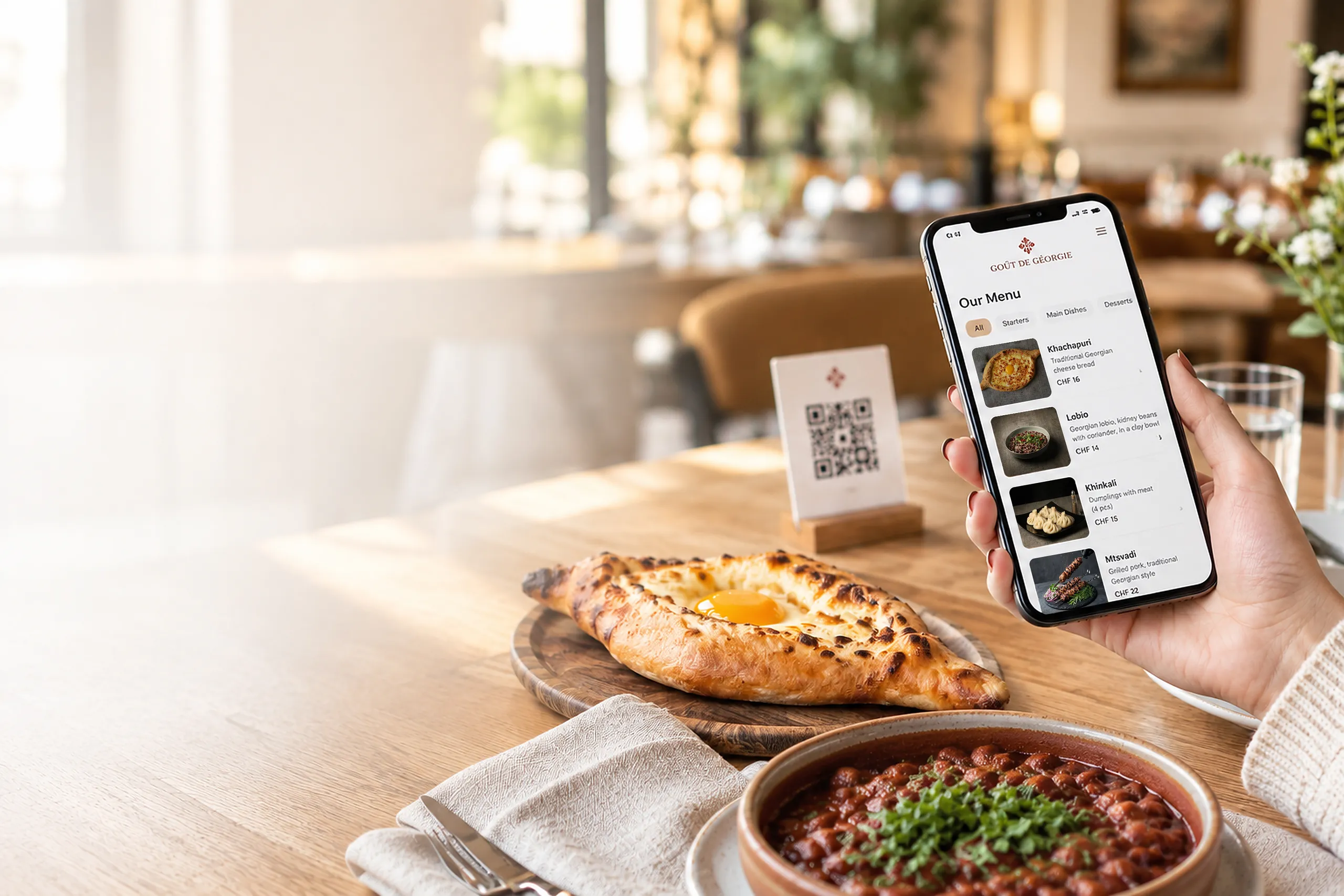Gast liest das QR-Menü von Goût de Géorgie auf dem Smartphone, mit Khachapuri, Lobio und QR-Aufsteller auf dem Tisch